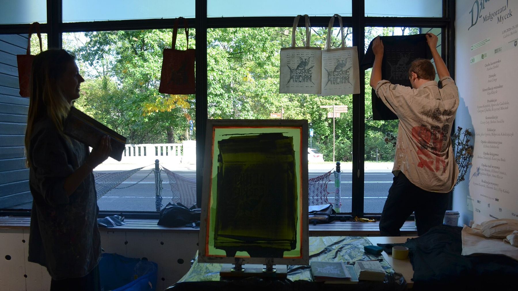 Silhouetted printmakers at a window-lit studio with a silkscreen frame and printed tote bags