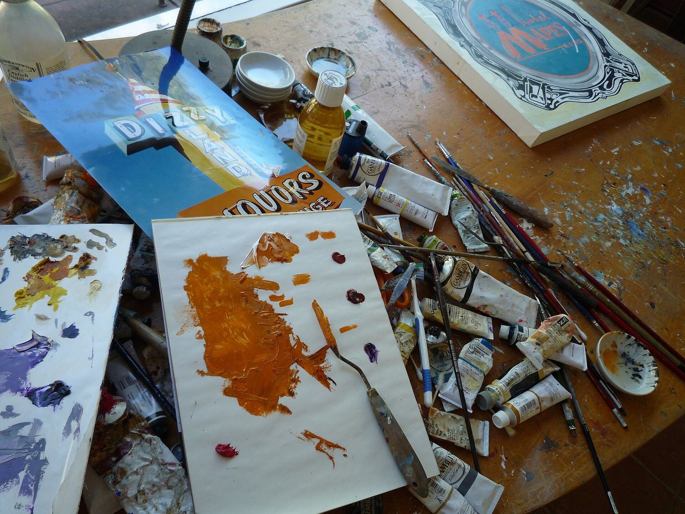 Paint-covered art desk with brushes, tubes and colourful sketches