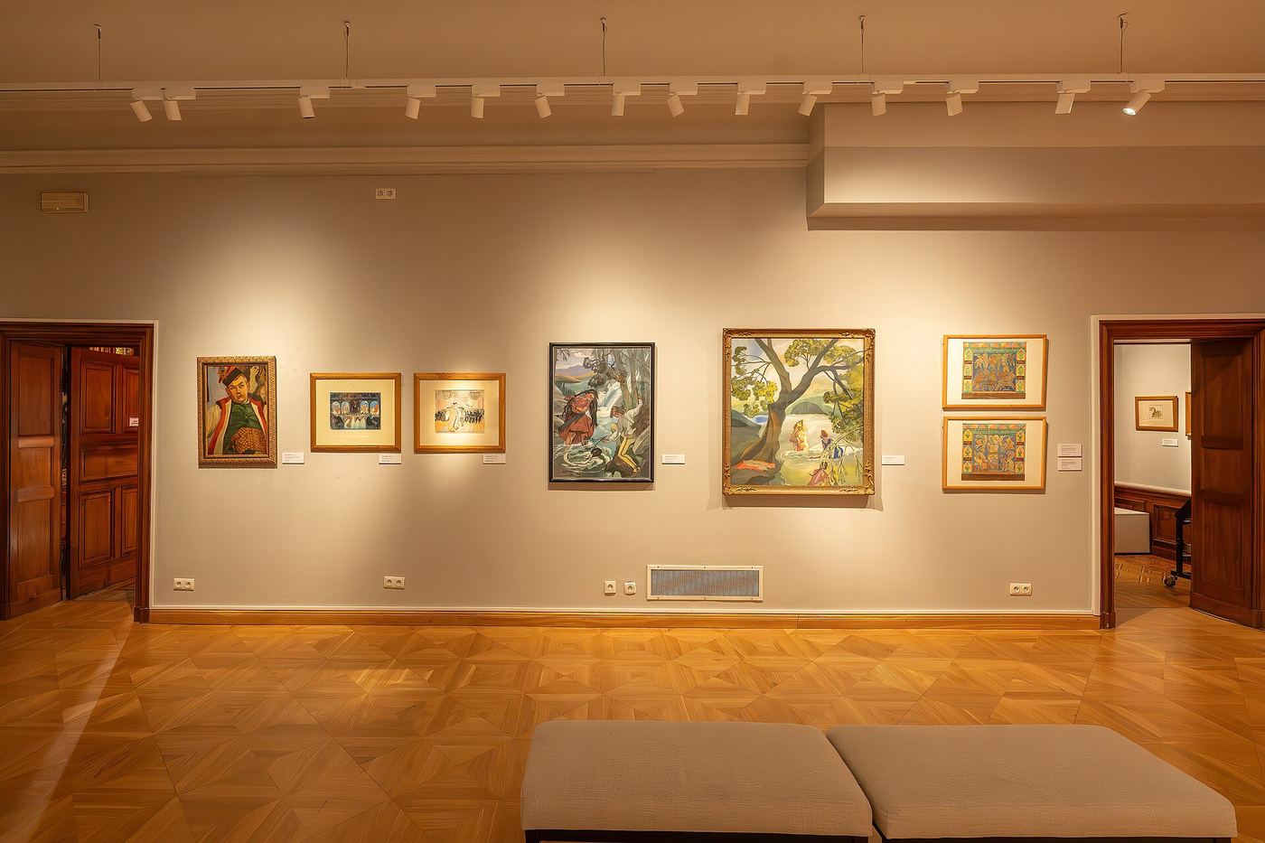 Warm-lit museum exhibition room with colourful framed paintings and a low bench