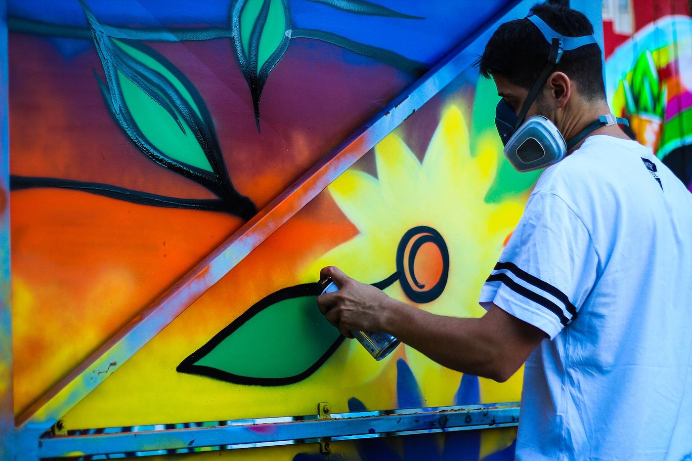 Artist in a respirator spray-painting a bright yellow flower on a mural