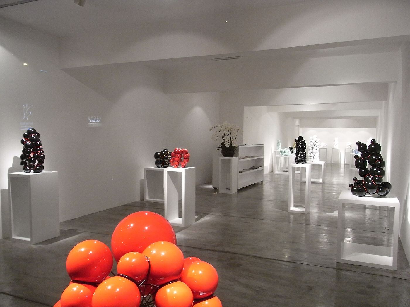 Bright white gallery with sculptures on plinths and orange spheres in the foreground